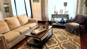 Un salón con un sofá beige, una mesa de centro con libros y objetos decorativos, una alfombra estampada y una mesita de noche con una lámpara y adornos.