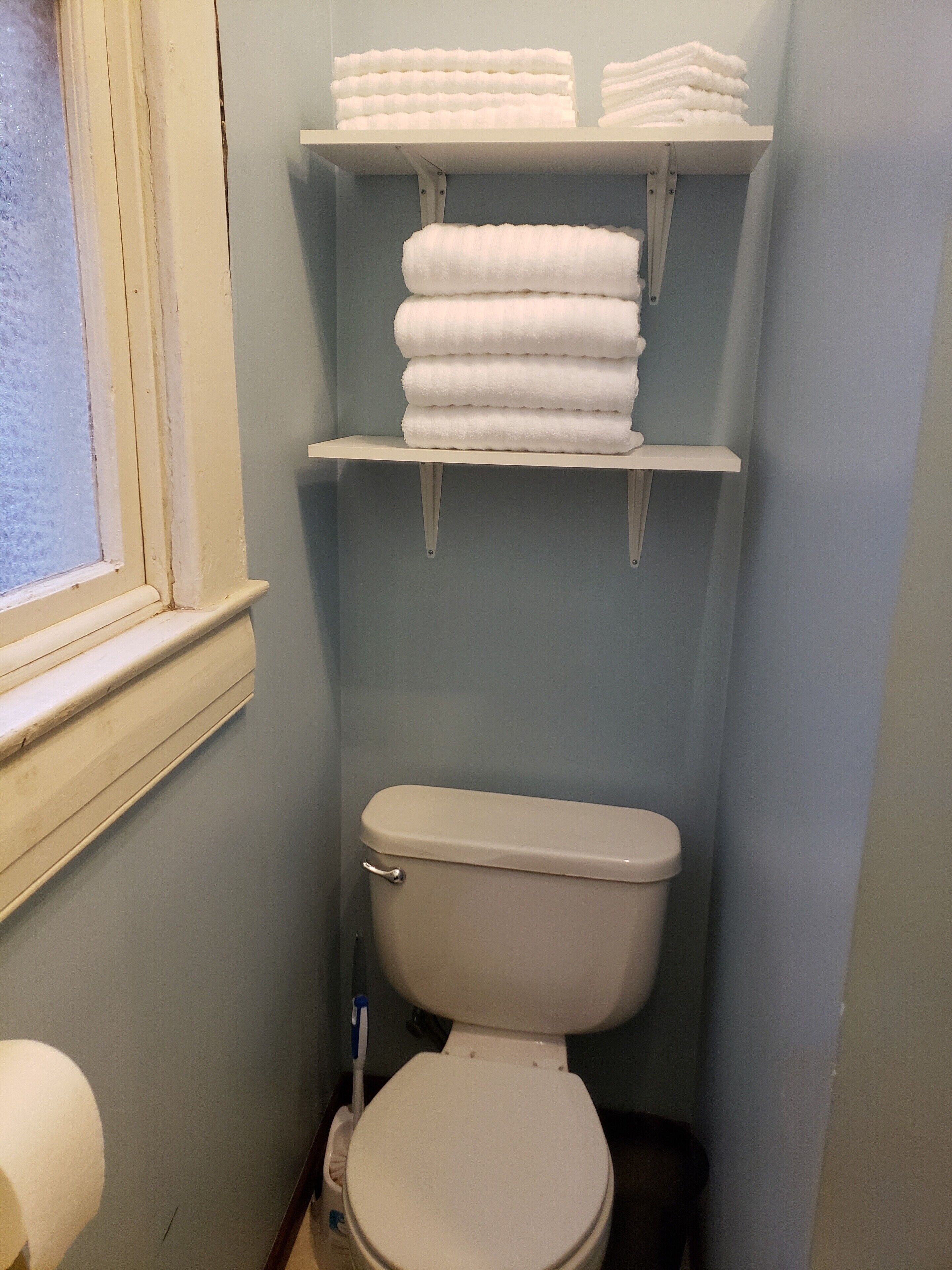 Une salle de bain avec des toilettes, une fenêtre et des étagères où sont pliées des serviettes.