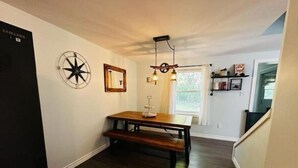 A dining area with a wooden table and bench, a wall clock, a decorative light fixture, and a window with curtains.