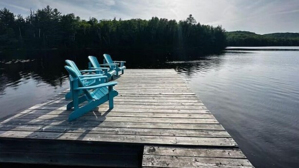 A wooden dock with blue Adirondack chairs facing a calm lake, surrounded by dense forest.