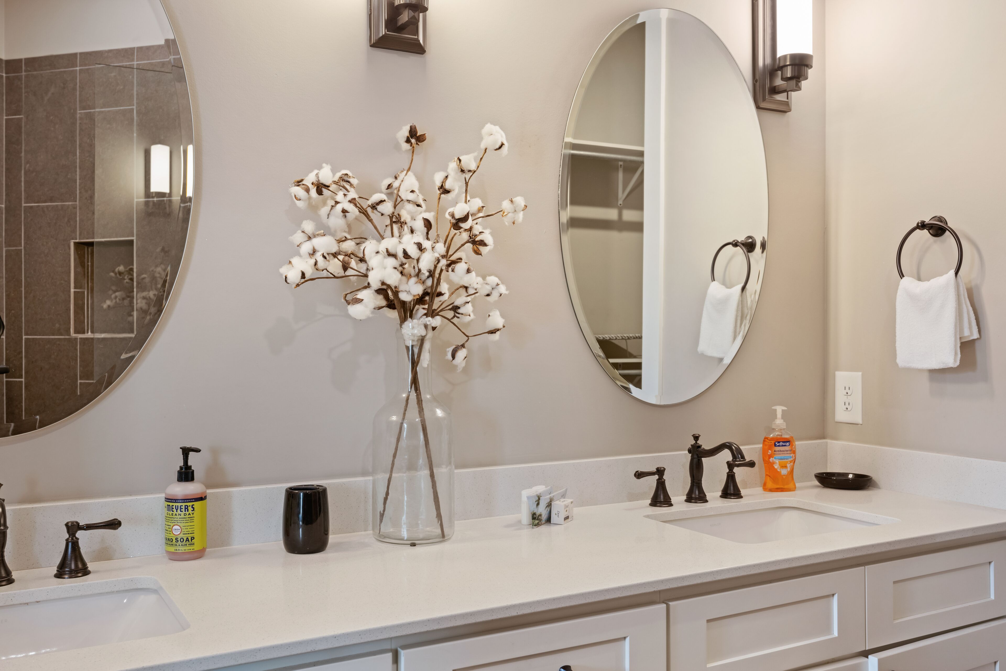 Une salle de bain avec un lavabo blanc, deux miroirs, un vase contenant des fleurs séchées, un distributeur de savon et un porte-serviettes.