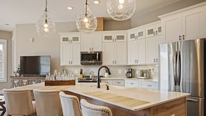 A modern kitchen with white cabinets, a stainless steel refrigerator, a large island with bar stools, and pendant lighting.