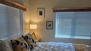 A bedroom with a large bed, two windows with blinds, a bedside lamp, and two framed pictures on the wall. - Tiny Farmhouse | CDA, Silverwood & Lake Nearby (Coeur d'Alene)