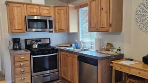 A kitchen with wooden cabinets, stainless steel appliances, a coffee maker, and a small dining area with a stool. - Tiny Farmhouse | CDA, Silverwood & Lake Nearby (Coeur d'Alene)