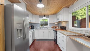 A modern kitchen with white cabinets, a stainless steel refrigerator, wooden ceiling, and a window with a view of greenery. - Couples Retreat! Every Season-Luxury, River Front, Hot tub and outdoor Shower (Glide)