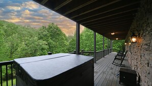 A covered outdoor deck with a hot tub, wooden railings, stone walls, and outdoor lighting. - Two Homes in One! Bear Hollow is the Perfect Luxury Escape for Large Gatherings! (Sevierville)