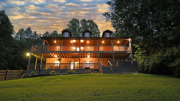 A two-story wooden cabin with a balcony, surrounded by a grassy yard and trees. - Two Homes in One! Bear Hollow is the Perfect Luxury Escape for Large Gatherings! (Sevierville)