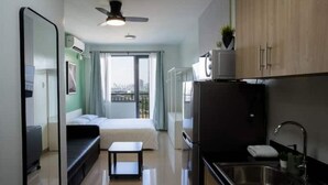 Een knus hotelkamer met een bed, een klein tafeltje, een kitchenette met een koelkast, en een raam met uitzicht op de stad.