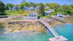 A coastal property with a wooden dock extending into clear waters, surrounded by lush greenery and multiple houses. - Linekin Lodge | Oceanfront | 3BDR| Deep Water Dock| Beach (Boothbay Harbor)