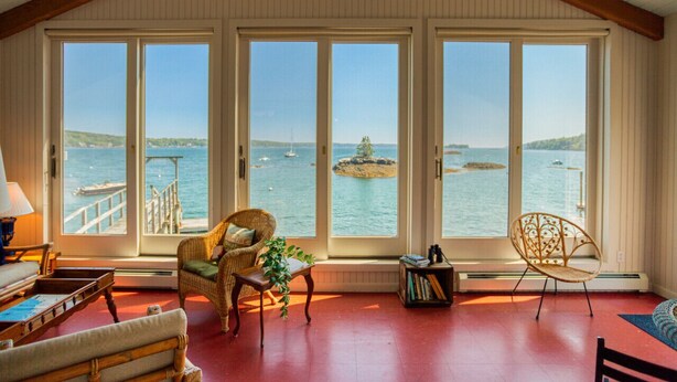 A cozy room with large windows overlooking a body of water, featuring wicker furniture and a ceiling fan. - Linekin Lodge | Oceanfront | 3BDR| Deep Water Dock| Beach (Boothbay Harbor)