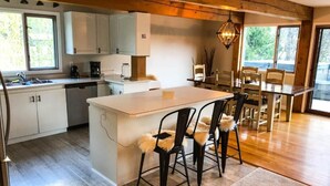 A modern kitchen with white cabinets, a central island with bar stools, and a dining area with a table and chairs. - Bearvalley Guesthouse (Stewart)