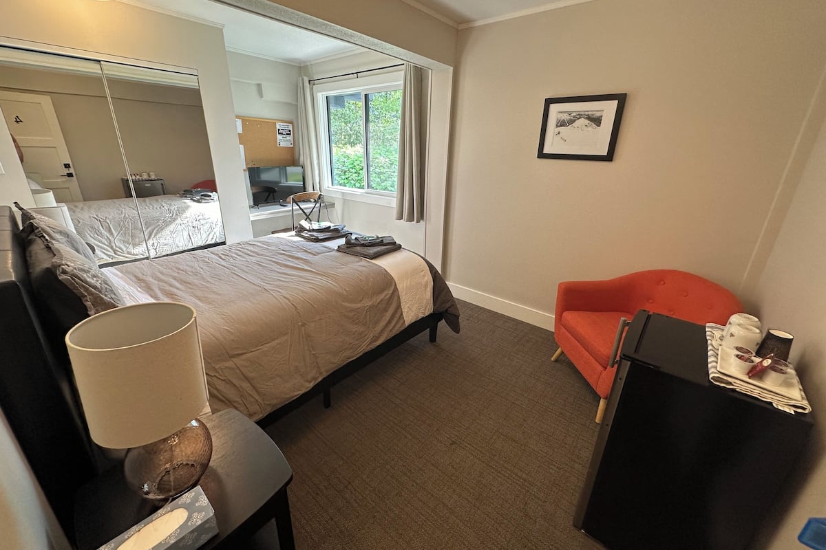 A hotel room with a bed, a desk, a chair, a window with a view of greenery, and a framed picture on the wall.