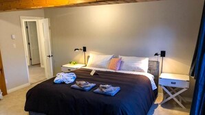 A neatly made bed with a black bedspread and multiple pillows, two bedside tables with lamps, and a door leading to another room. - Bearvalley Guesthouse (Stewart)