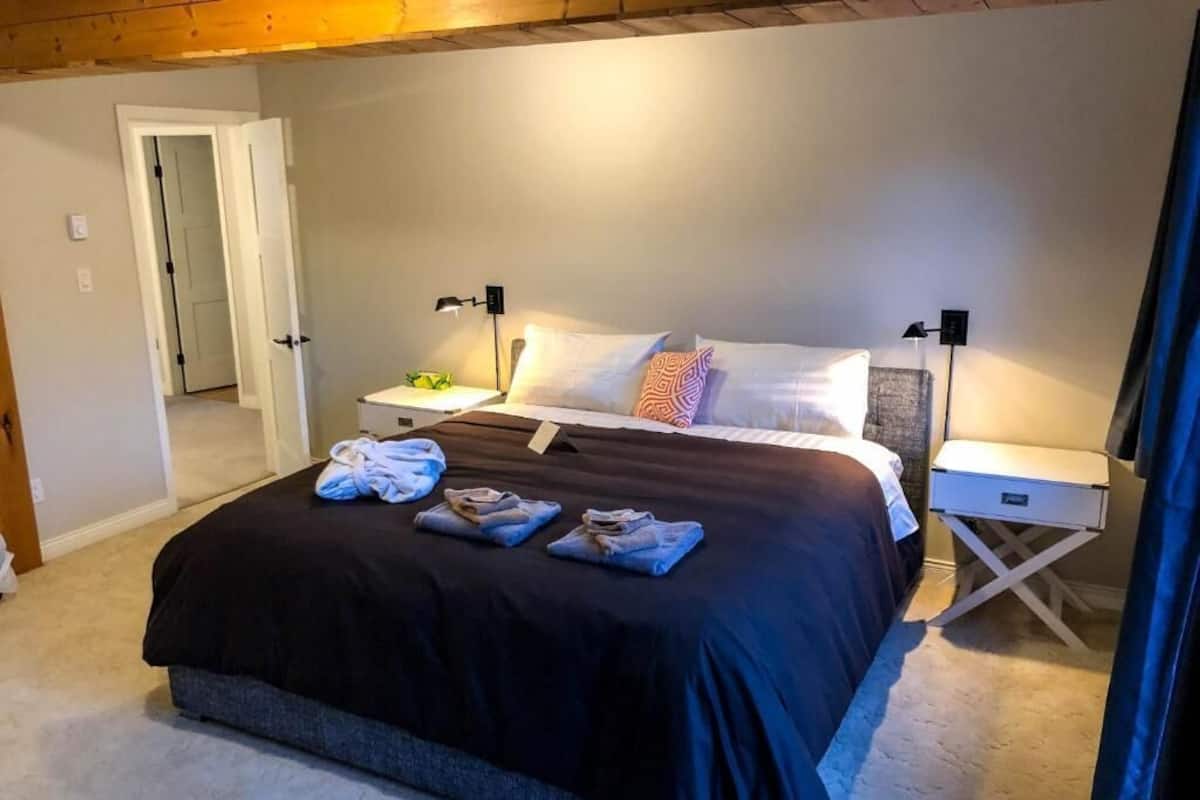 A neatly made bed with a black bedspread and multiple pillows, two bedside tables with lamps, and a door leading to another room.