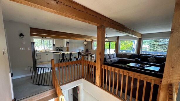 A spacious living area with a wooden staircase, a kitchen in the background, and large windows offering views of greenery. - Bearvalley Guesthouse (Stewart)