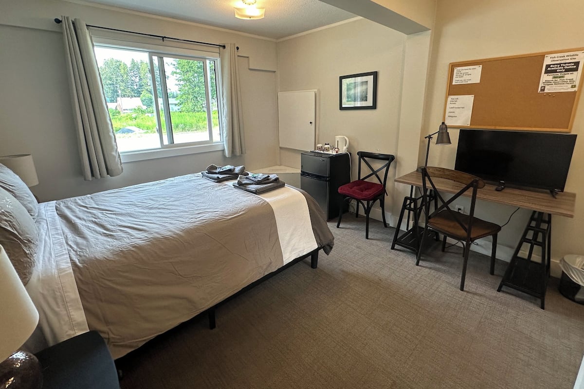A hotel room with a bed, a desk with a computer, a chair, a TV, and a bulletin board.