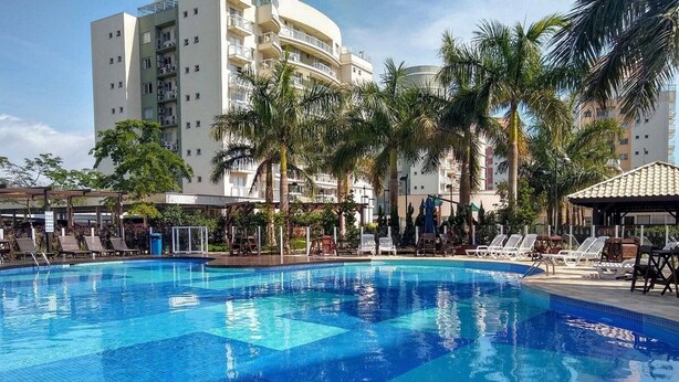 A swimming pool with lounge chairs, palm trees, and a multi-story building in the background. - Nautilus Paraíso Tropical - Vista ao Mar (Penha)