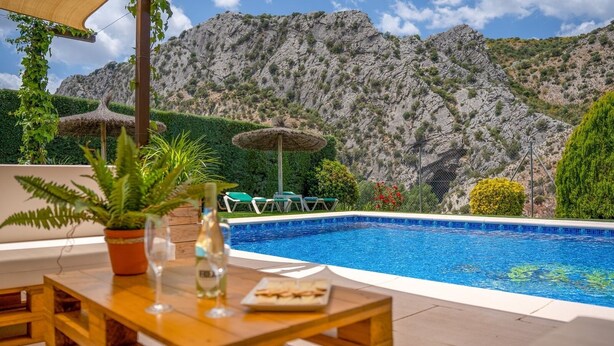 A pool area with a mountain backdrop, a table with a potted plant, and lounge chairs. - Casitas Cueva del Gato Benaoján by Ruralidays (Málaga)