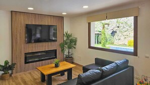 A modern living room with a flat-screen TV, a fireplace, a wooden table, a sofa, and a large window offering a view of greenery. - Casitas Cueva del Gato Benaoján by Ruralidays (Málaga)