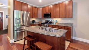 A modern kitchen with wooden cabinets, stainless steel appliances, granite countertops, and a central island with stools. - Riverfront Condo w/ Hot Tub | River Swimming Spot! (Lincoln)