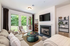 A living room with a fireplace, a television, a bookshelf, and a view of the outdoors.