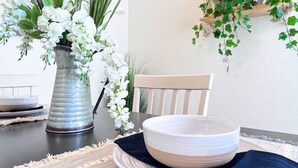 Una mesa de comedor con un plato y un bol blanco, un florero de metal con flores blancas y un colgador de macramé en la pared.