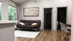 A modern living room with a black leather sofa, a white rug, and a dining area with a grey chair and a wooden table.