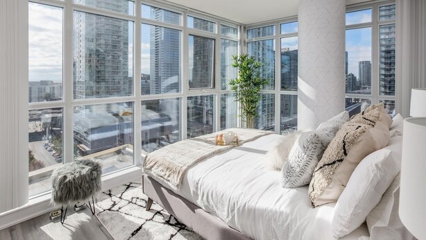 A modern bedroom with a large window, a bed with white bedding, a fluffy stool, and a potted plant.