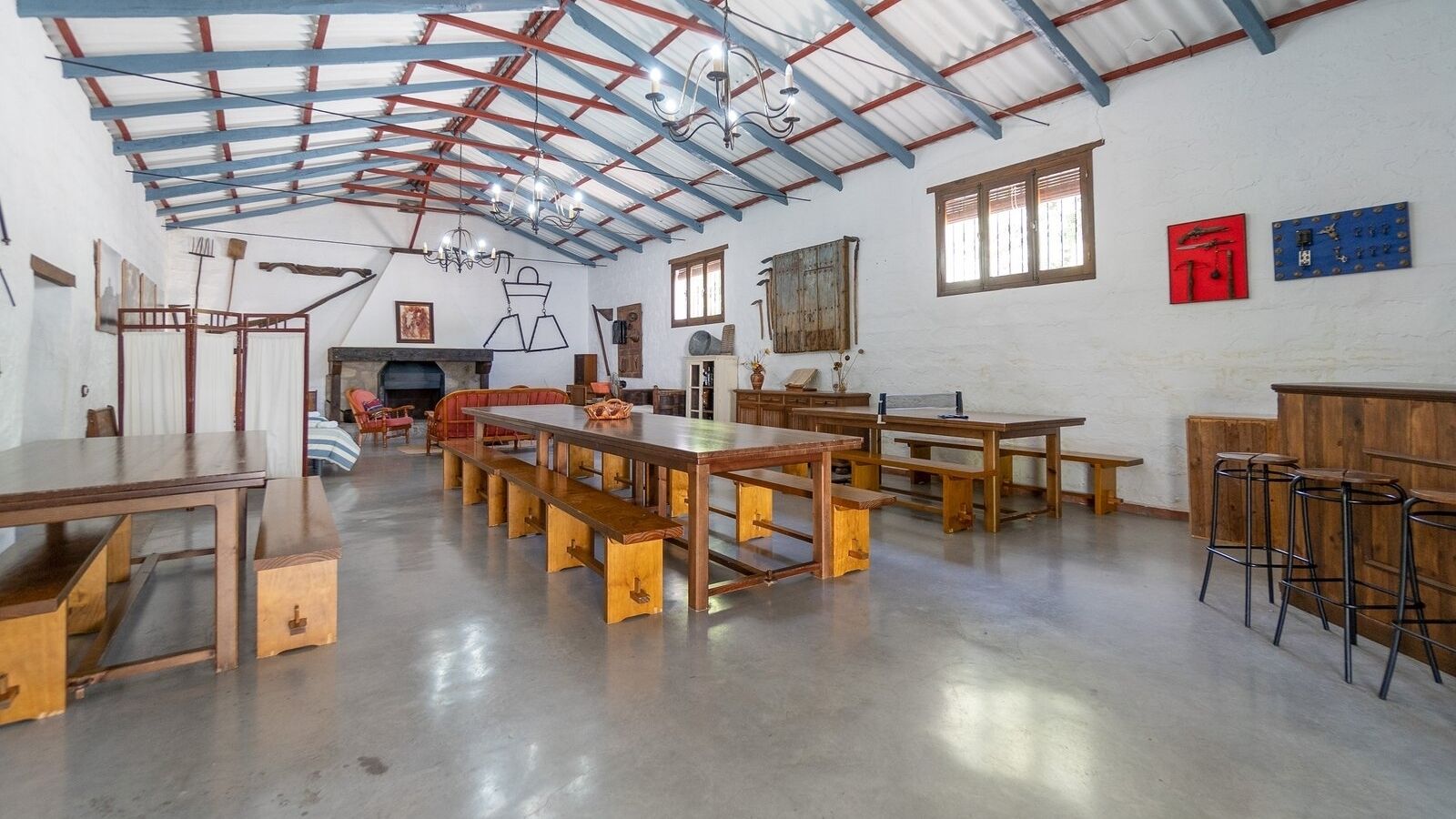 Amplia habitación con techo alto, mesas y bancos de madera, y una zona de bar con taburetes.