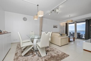 A modern living room with a glass dining table, white chairs, a beige sofa, and a view of the sea through large windows. - C te D Atlantique by Homie (Santa Cruz)