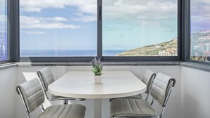 Ein modernes Zimmer mit einem runden Tisch und vier Stühlen, große Fenster mit Blick auf das Meer und eine hügelige Landschaft.