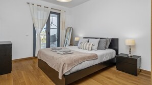 Ein Schlafzimmer mit Bett, Nachttischlampen, einem Nachttisch, einem Fenster mit Vorhängen und Blick auf Gebäude.