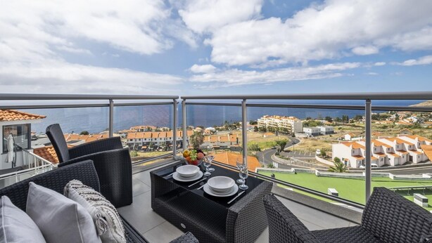 Ein Balkon mit Korbmöbeln, einem für zwei Personen gedeckten Esstisch und einem schönen Ausblick auf eine Küstenstadt.