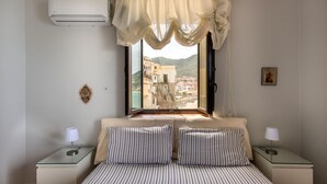 A bedroom with a bed, two bedside tables, a window with curtains, an air conditioner, and a view of buildings. - Meravigghia Apartment by Wonderful Italy (Cefalù)