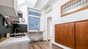 Una cocina moderna con encimera blanca, pileta de acero inoxidable y dos taburetes de bar. Se observa un ventanal grande sobre la pileta, y un armario de madera a la derecha.