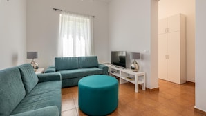 A modern living room with a teal sofa, a matching ottoman, a TV on a white stand, and a wardrobe.