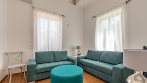 A living room with two teal sofas, a teal ottoman, a small white chair, and a table with a lamp.