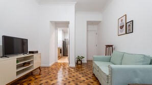 A living room with a TV, a light blue sofa, wooden parquet flooring, and a kitchen visible through an open doorway. - 5 Minutes From Leblon Beach Ap102 (Rio de Janeiro)