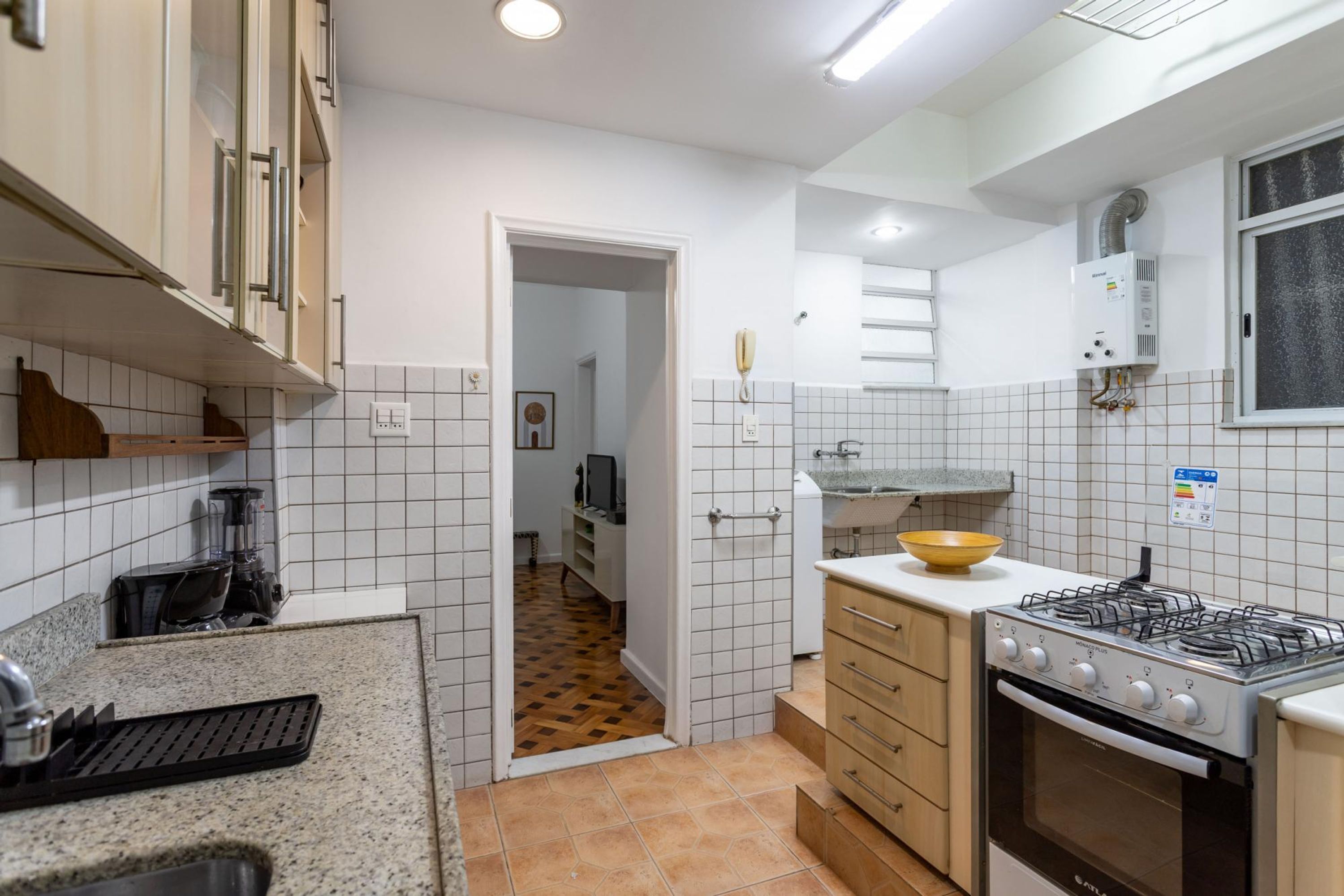 A kitchen with white tiled walls, a gas stove, a built-in oven, and a countertop with a kettle and a bowl.