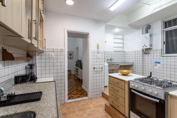 A kitchen with white tiled walls, a gas stove, a built-in oven, and a countertop with a kettle and a bowl.