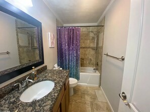 Een badkamer met een wastafel met granieten blad, toilet, bad en douche met paars gordijn.