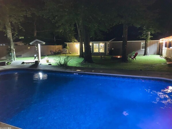 A backyard with a swimming pool, a covered patio, and a small house with a lit window.