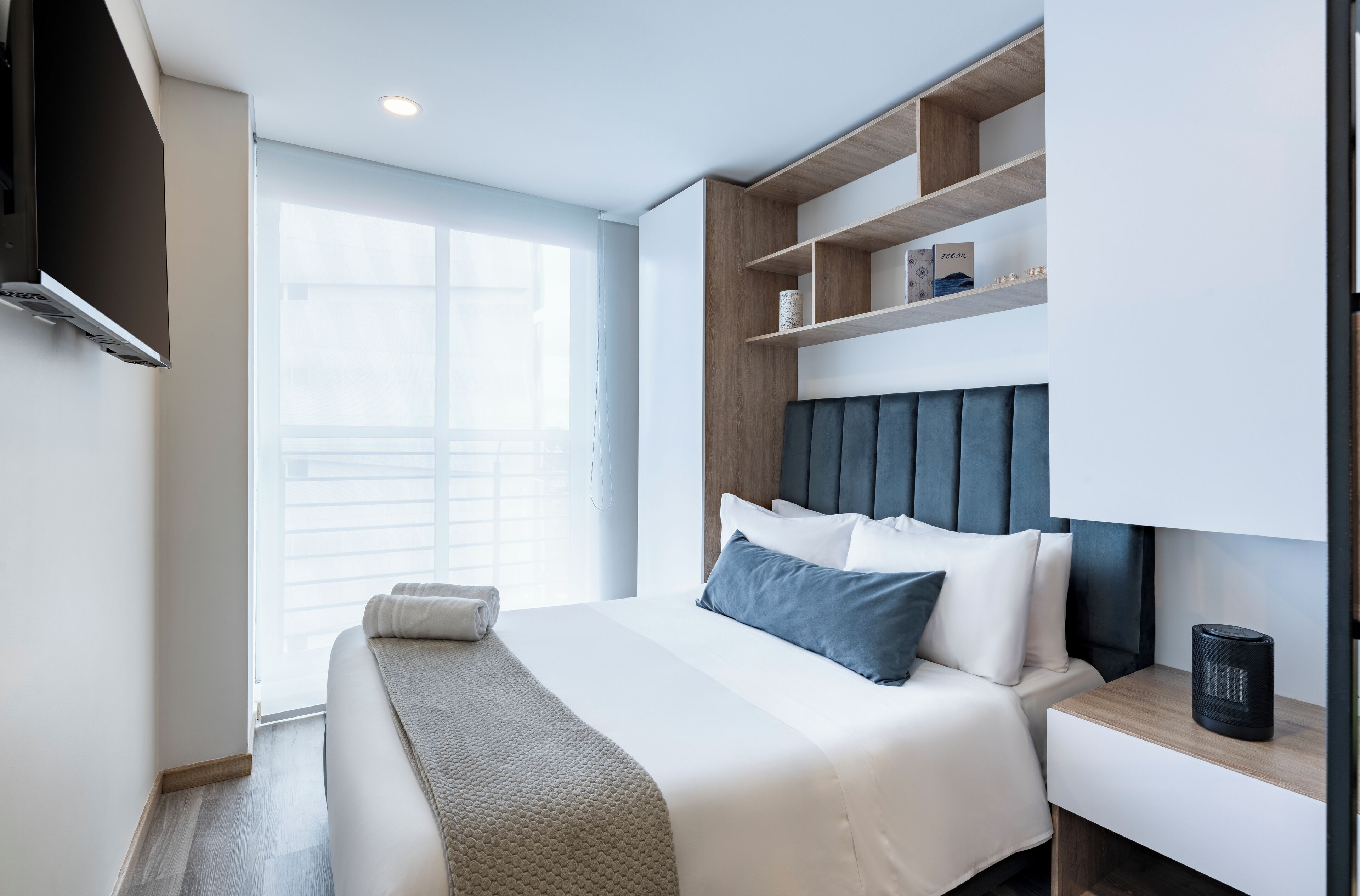 A modern hotel room with a large bed, a bedside table, a built-in shelf unit, and a window with blinds.