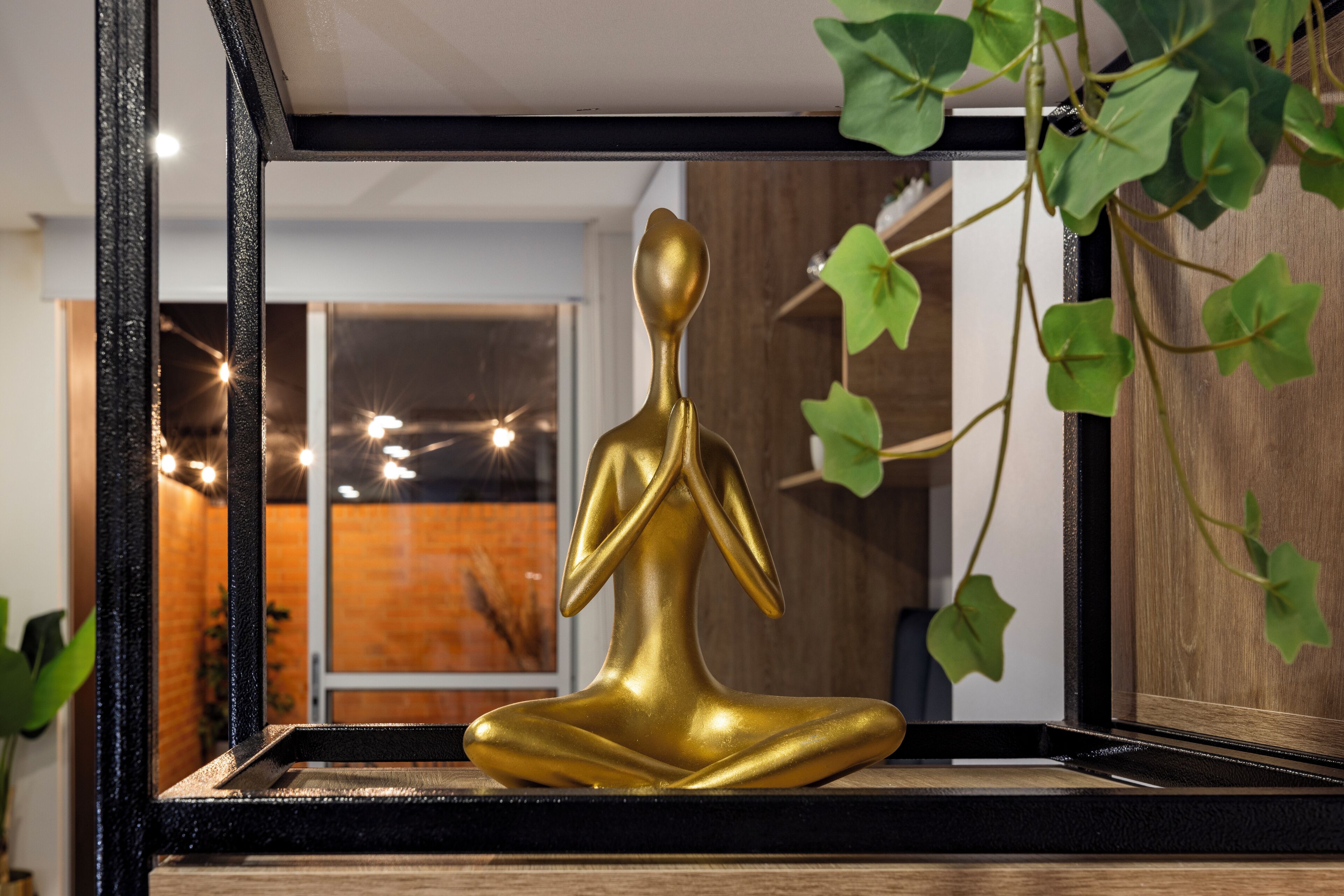A golden statue of a seated figure on a wooden platform, surrounded by greenery and a black-framed mirror.