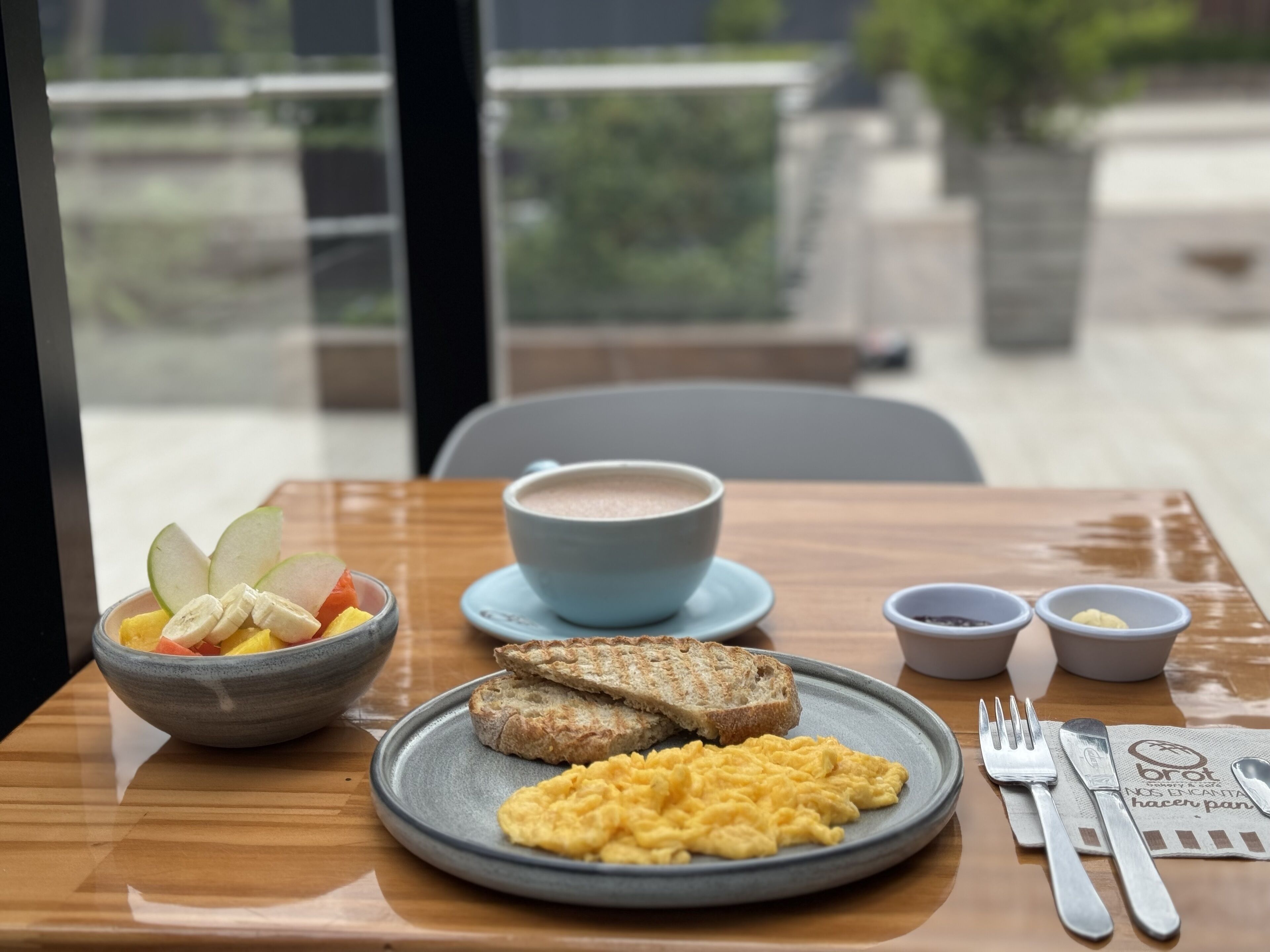 A breakfast setting with a bowl of fruit, scrambled eggs, toast, and a cup of coffee on a wooden table.