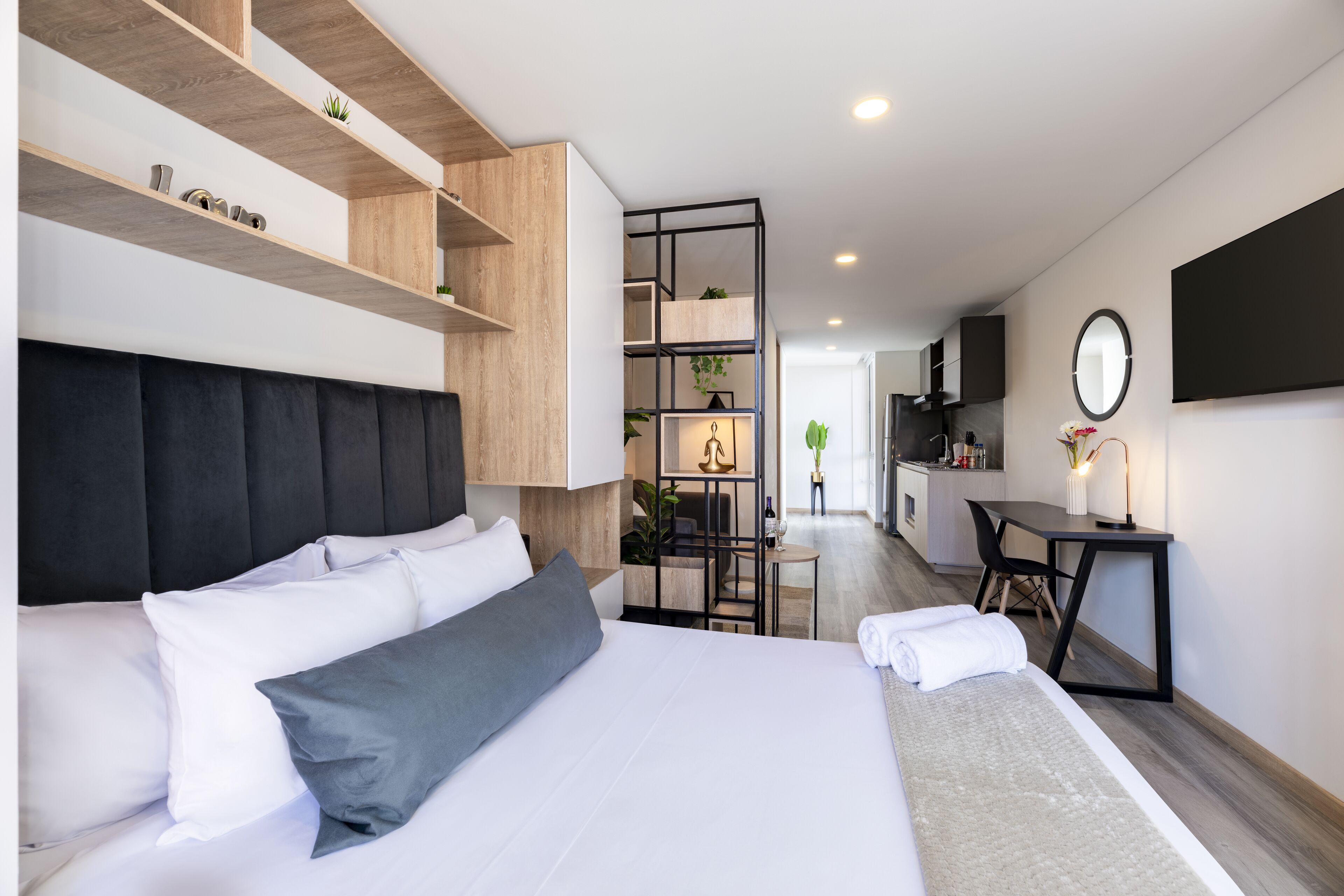 A modern hotel room with a large bed, a desk, and a kitchenette in the background.