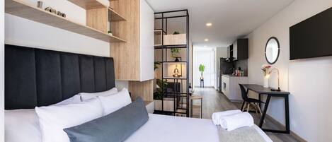 A modern hotel room with a large bed, a desk, and a kitchenette in the background.