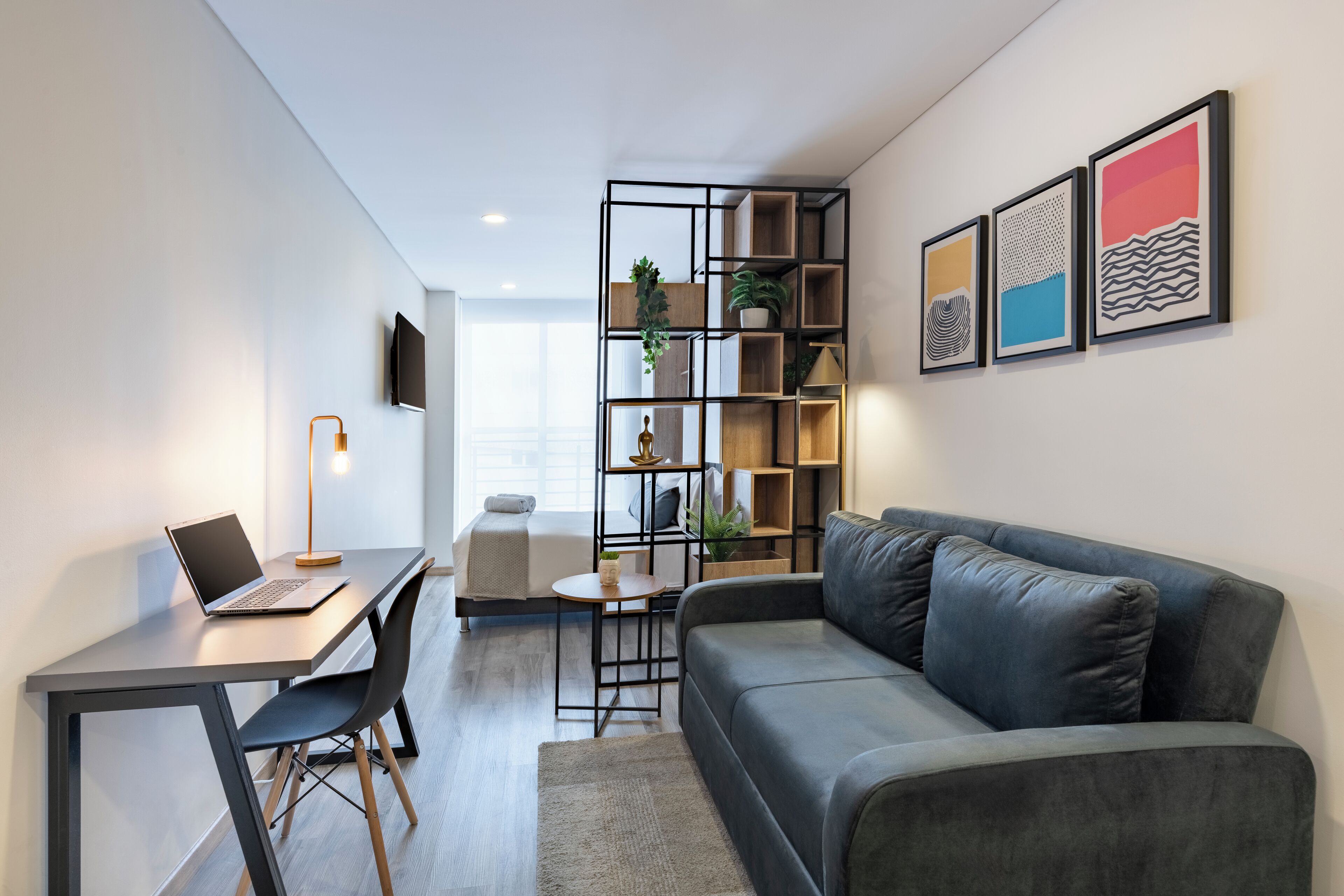 A modern living room with a grey sofa, a desk with a laptop, a small round table, and a bookshelf.