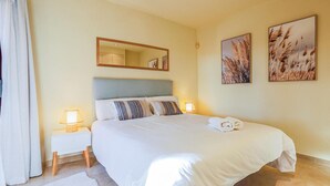 Ein ordentlich bezogenes Bett mit weißen Leinen und einem gefalteten Handtuch, ein Nachttisch mit Lampe, ein Spiegel über dem Bett und zwei an der Wand hängende Bilder.