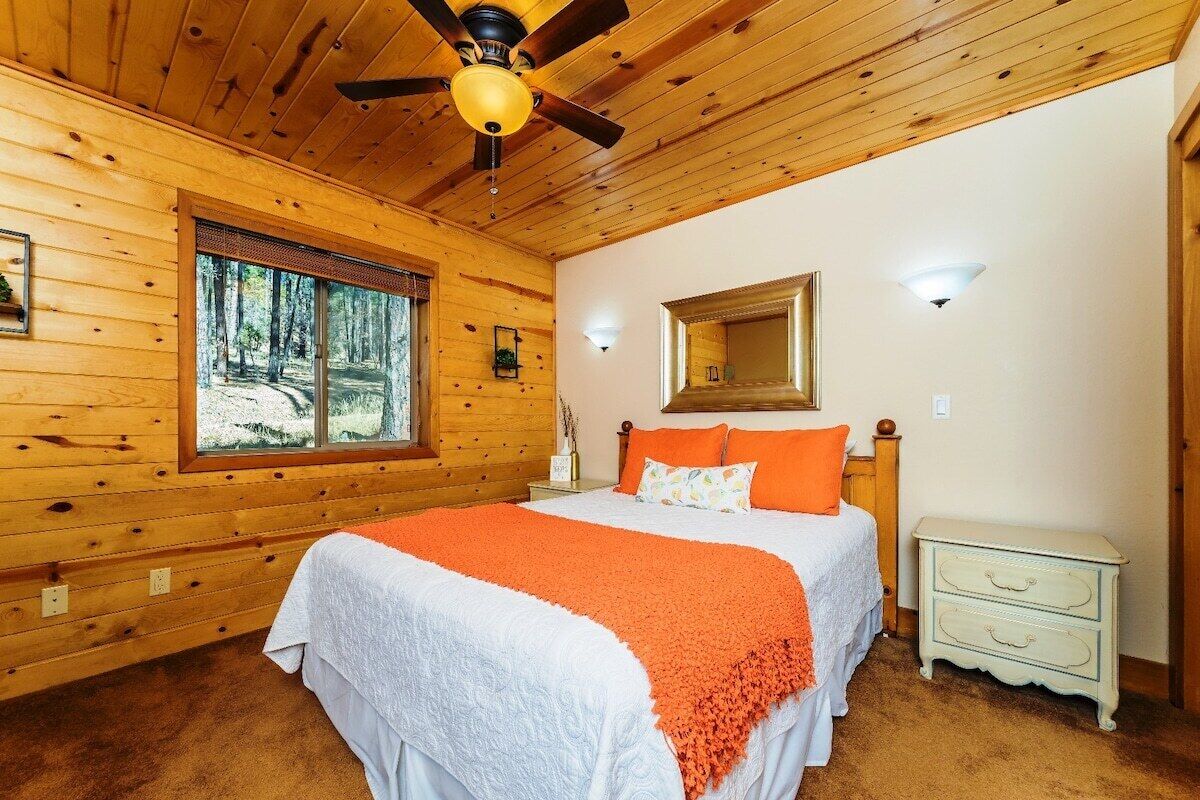 A wooden cabin bedroom with a large bed, a ceiling fan, a window with a forest view, and a nightstand with a lamp.